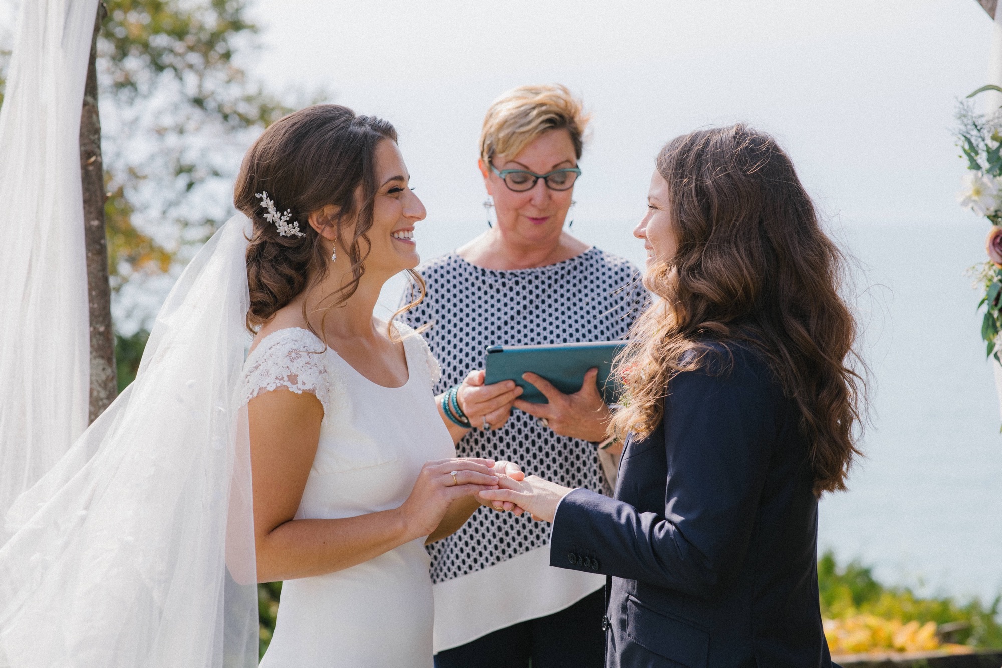 Taylor and Nicole Johnston wearing a wedding dress and suit hold hands at their outdoor wedding ceremony with officiant Sherry Murphy standing in the middle of them. 