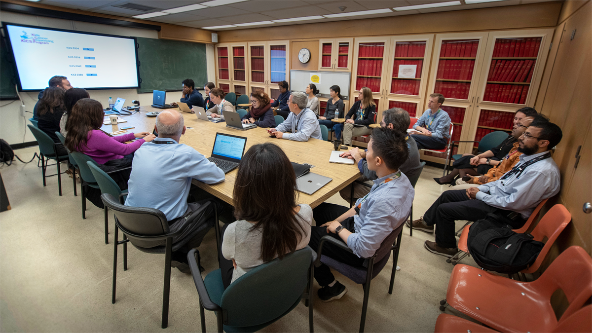 A group of about 20 people sitting around a table in a boardroom. Many of them have their laptops open or looking toward a large screen at the front of the room that is displaying a presentation slide.