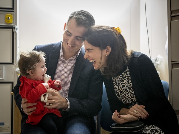 Baby sits on her father's lap. Her mother leans towards her. They are all smiling.