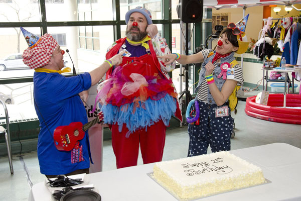 Three clowns stand together with a birthday cake in front of them.