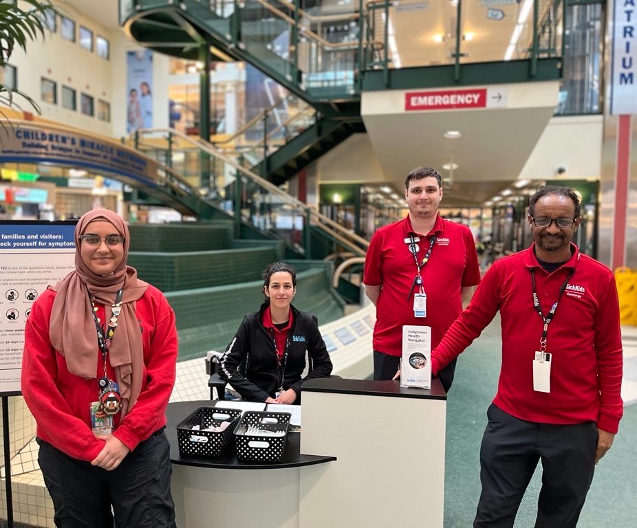 Four members of the hospital concierge team stand and sit at an information desk in the hospital atrium. The team is available to welcome visitors and help people find their way through the hospital.