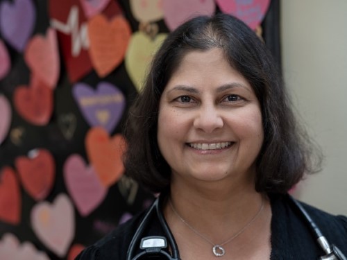 Wearing a black shirt and stethoscope, Dr. Anne Dipchand smiles in front of colourful paper hearts on the wall.