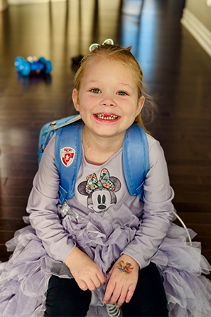 A young girl smiles at the camera, her backpack has tubes to provide her with nutrition.