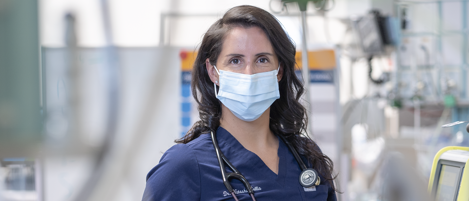 Dr. Natasha Collia in a mask with a stethoscope around her neck. There is a variety of hospital equipment out of focus in the background.