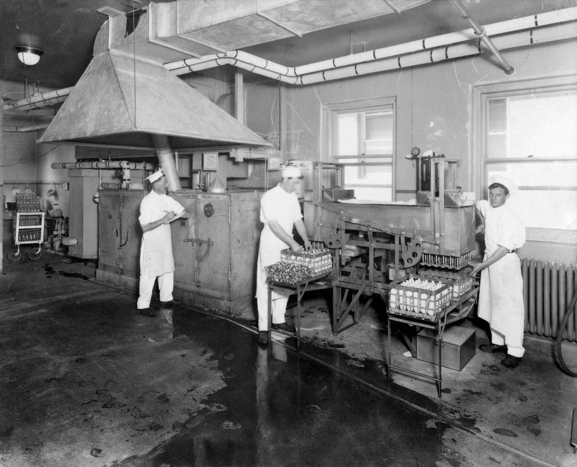 SickKids milk pasteurization plant from 1909 showing three workers in white uniforms processing bottles of milk in an industrial setting.