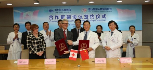 Two hospital representatives shake hands and hold signed documents at a cooperation agreement ceremony between SickKids Hospital and a Chinese medical institution. Canadian and Chinese flags are displayed, with medical staff in white coats witnessing the international healthcare partnership.