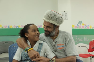 A smiling father with gray beard and knitted cap embraces his daughter who wears a school uniform with plaid skirt. They sit in a colorful classroom with painted flower decorations on the wall.