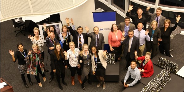 A group photo of staff members from SickKids Centre for Global Child Health. About twenty-five professionals in business attire wave at the camera in a modern office space, all wearing ID badges. The organization's banner is visible in the background.