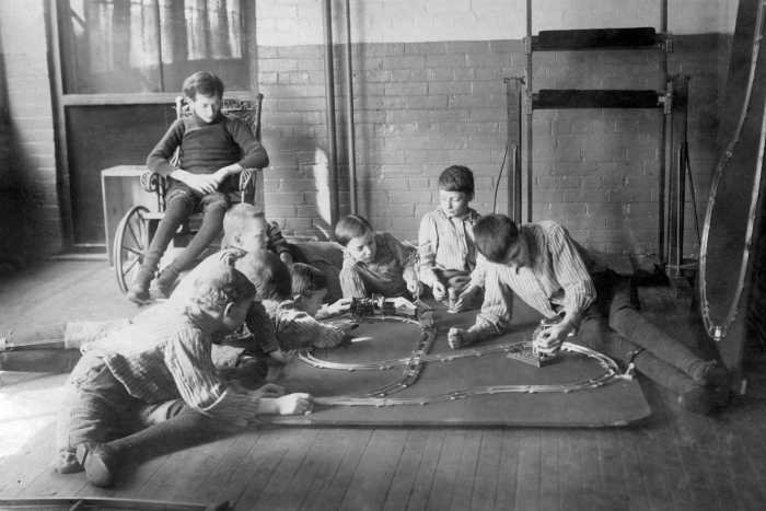 Vintage black and white photo of boys playing with toy train set while one sits in wheelchair in brick-walled room.