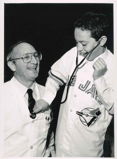 Black and white photo of doctor with young boy wearing Blue Jays jersey playing with stethoscope, both smiling.