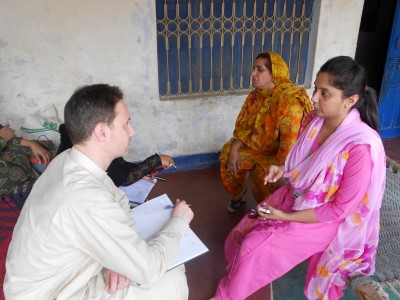 A healthcare worker or researcher in light clothing interviews two South Asian women wearing traditional clothing - one in orange/yellow and one in pink. He appears to be taking notes during what looks like a community health survey or field research in a simple indoor setting.