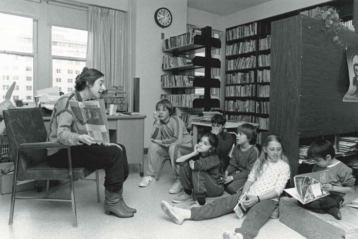 Black and white photo of woman reading to children sitting on floor in hospital library with bookshelves behind them.