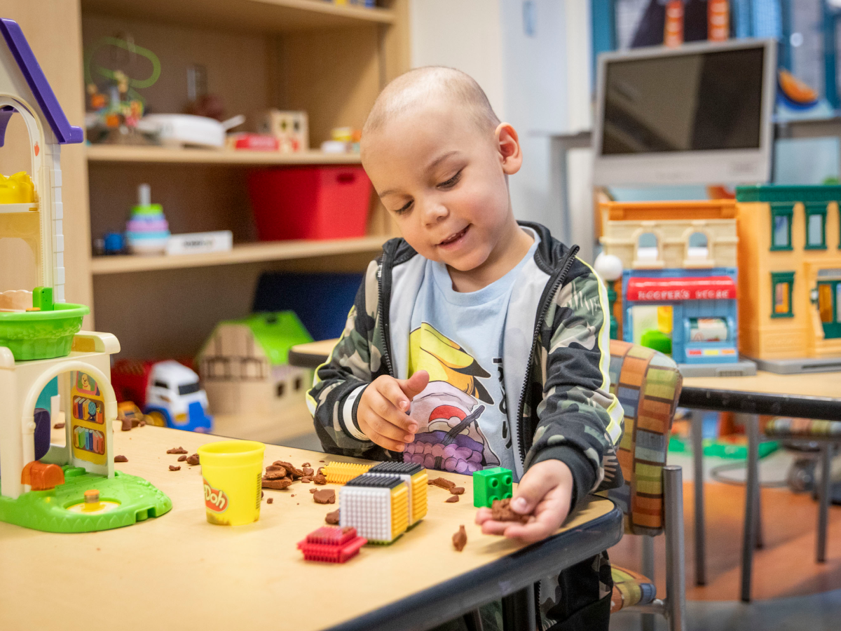 Smiling young kid playing with toys and Play-Doh in a hospital playroom surrounded by colorful educational toys and games.