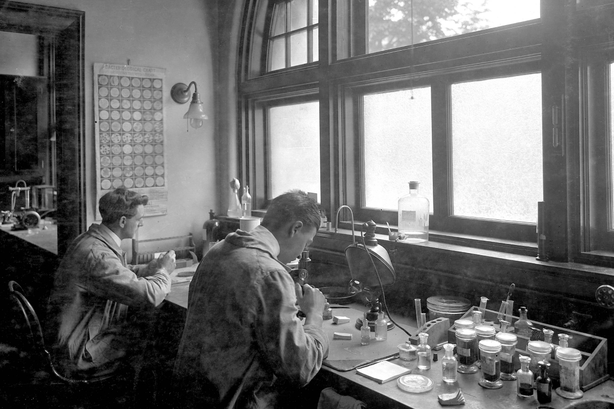 Black and white photograph of two scientists working at a laboratory bench with glassware and equipment. Large windows provide natural light. A periodic chart is visible on the wall.