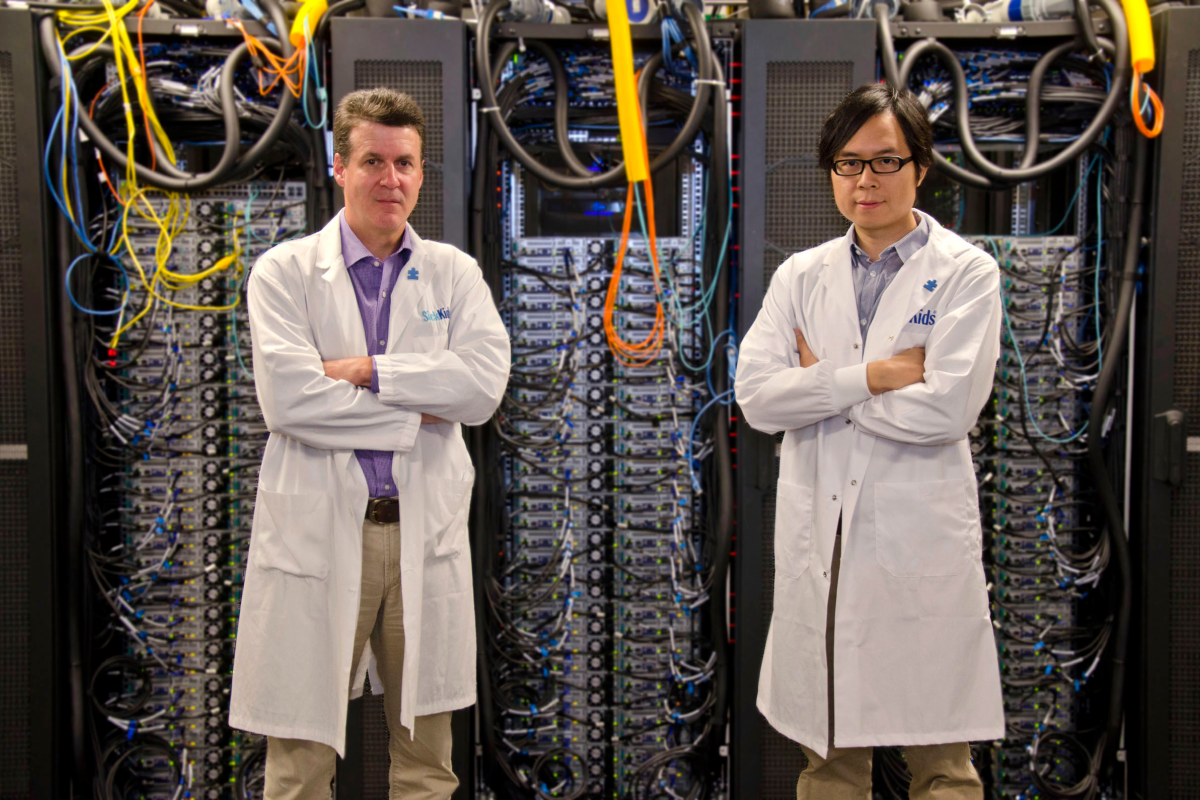 Two SickKids researchers in white lab coats stand in front of advanced computing server racks with colorful cables.