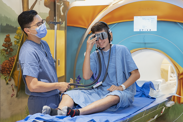 Teen boy looks into virtual reality goggles while seated on an MRI bench. A staff member stands by.