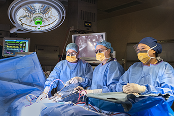 Three people dressed in medical gowns, gloves, caps and goggles stand at a surgical bed in an operating room.