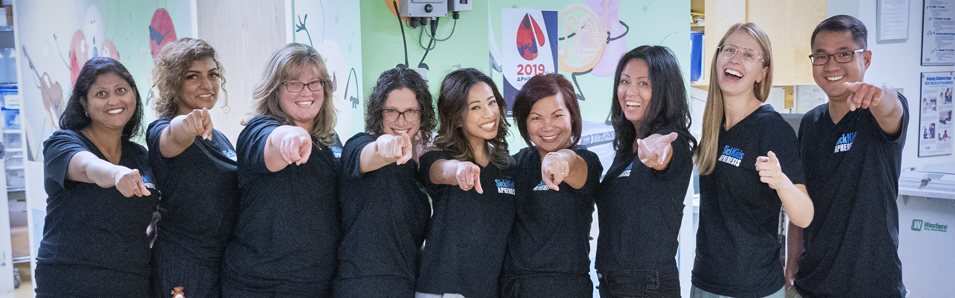 Nine people stand together smiling, pointing at the camera.