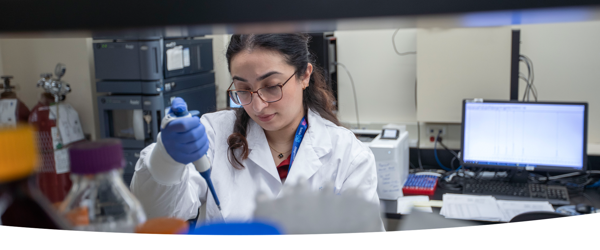 A lab professional wearing a lab coat and blue gloves using a large pipette. In the background is various lab equipment and a computer screen.