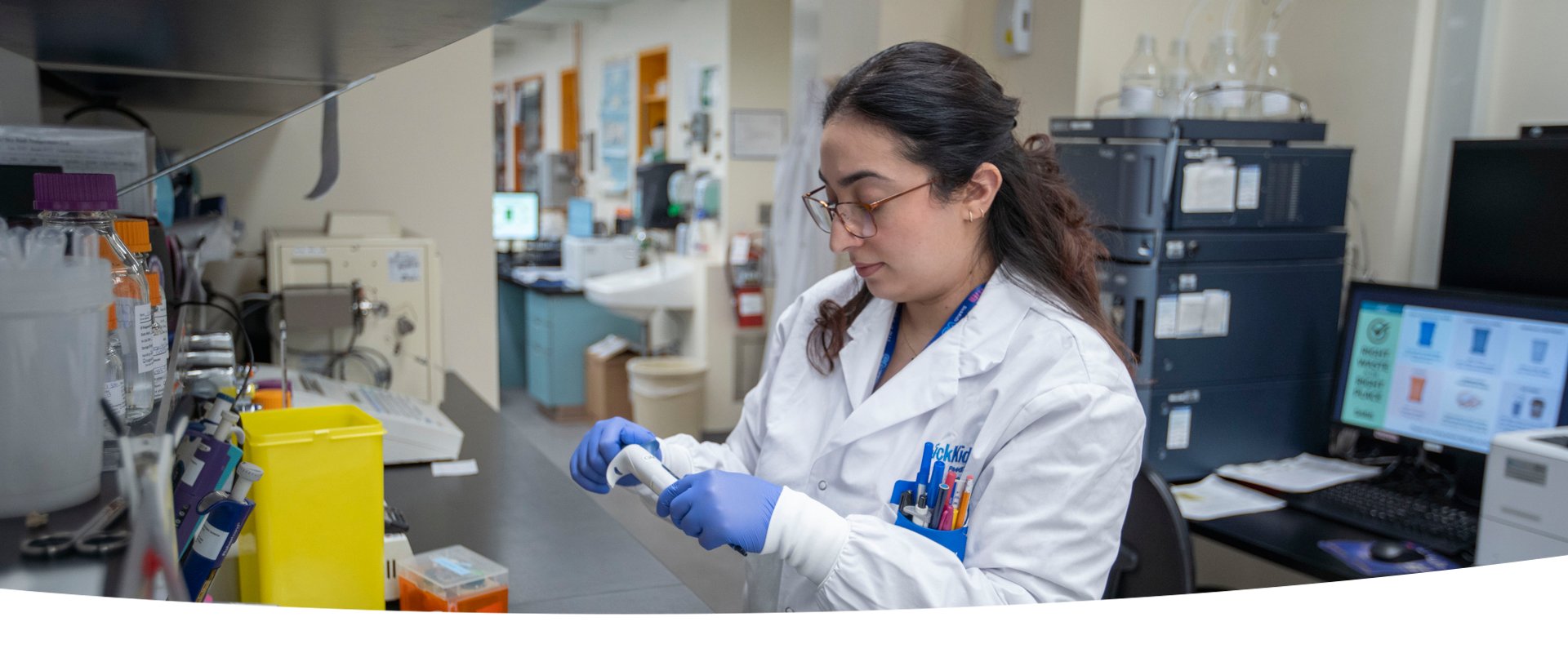 A lab professional wearing a lab coat and blue gloves. She is handling a piece of equipment at a lab bench.
