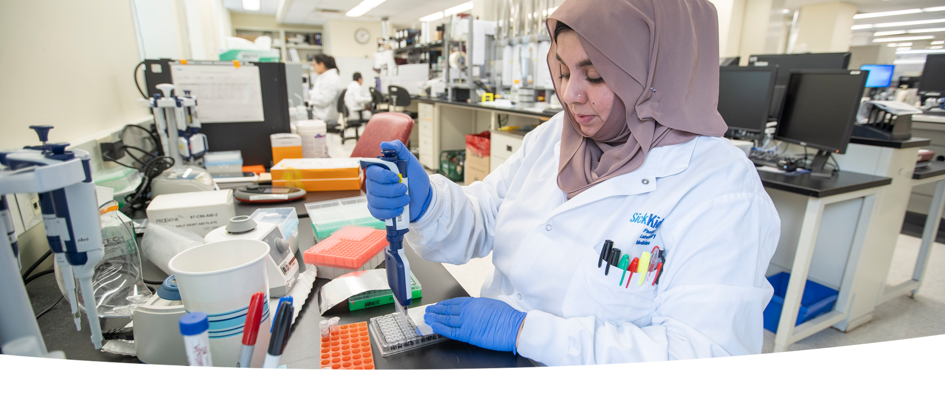 A laboratory professional pipetting into a multiwell plate at a lab bench. In the background, there are other lab professionals at work.