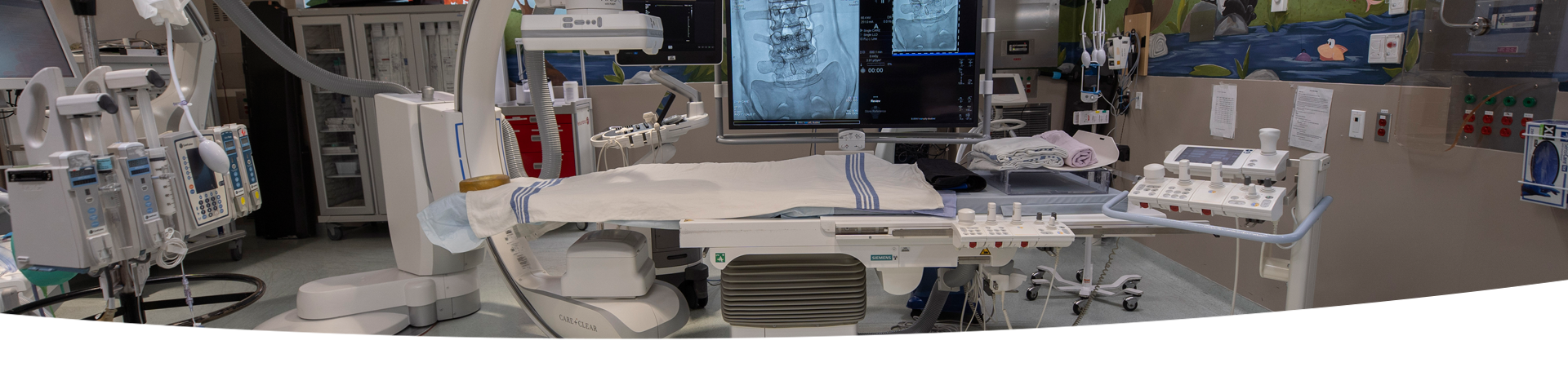 A room with a Single Plane Digital Angiography unit. There is a  variety of hospital equipment in the background.