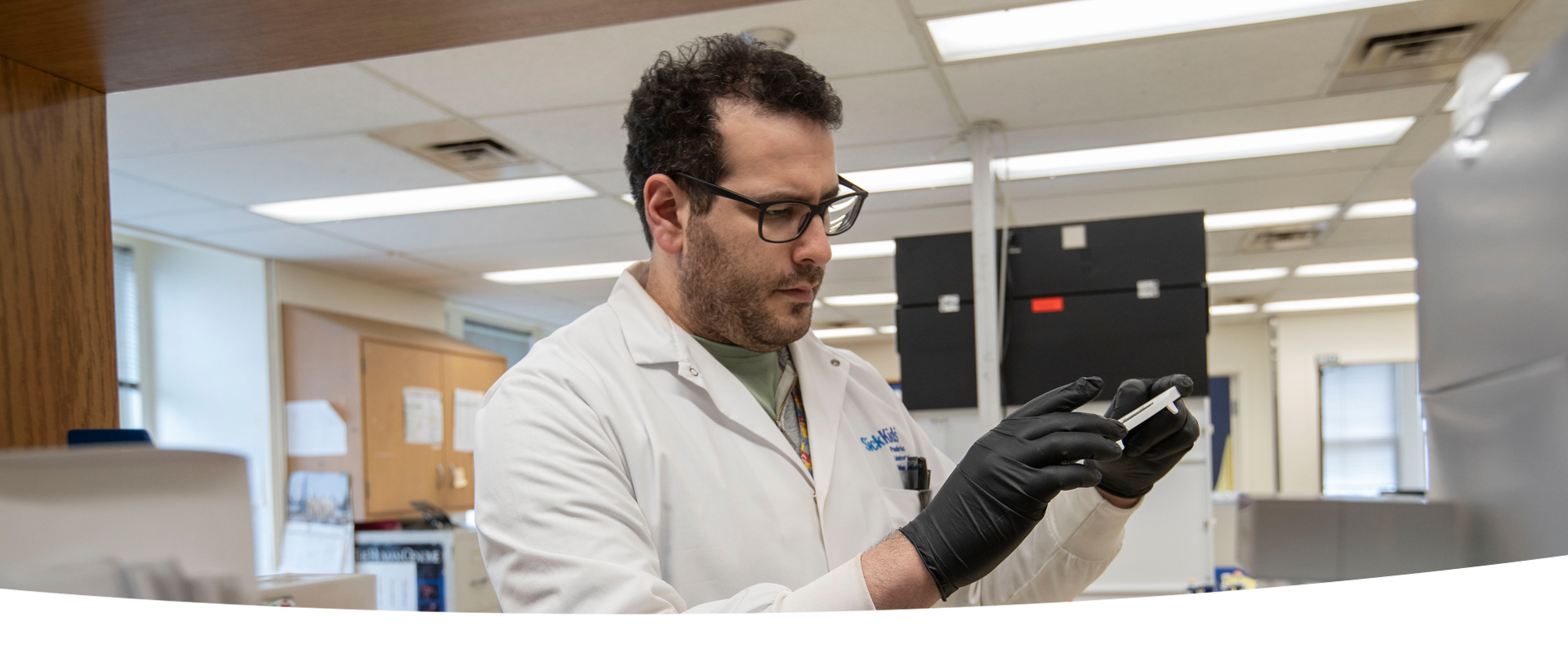 A lab professional wearing a lab coat and black gloves is examining a piece of equipment in his hands.