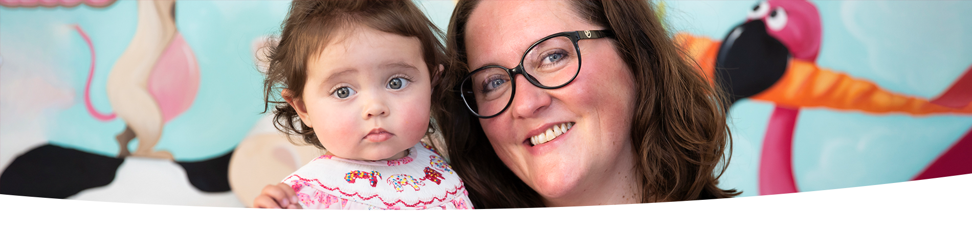 An infant and a woman in glasses pose together for a photo in front of a colourful mural. The mural has paintings of cartoon animals, including a cow, flamingo, and mouse.