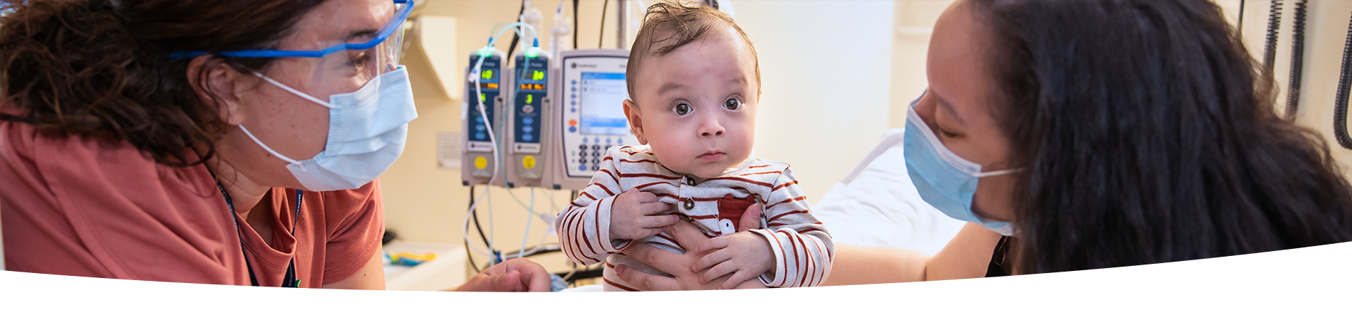 Two women hold a baby seated upright in a hospital bed. They both wear medical masks.