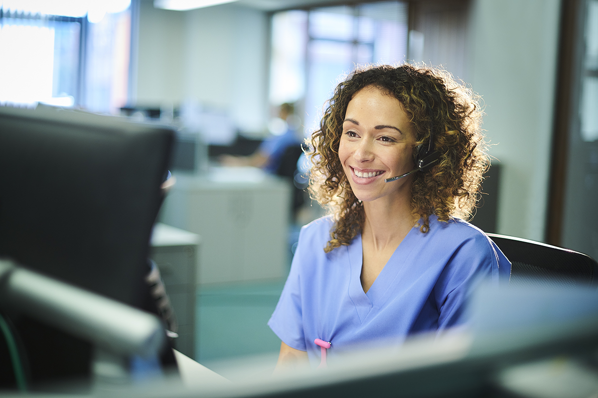 Woman wearing headset and hospital scrubs, seated at a desk.