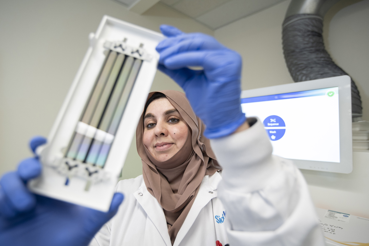 A laboratory professional wearing blue gloves and a white lab coat holds up a piece of laboratory equipment with thin, reflective strips. She appears focused, with a computer screen displaying sequencing-related information in the background.