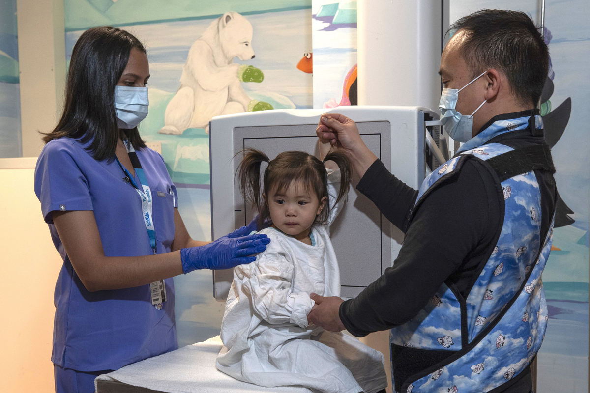 A child wearing a protective gown for X-rays while two staff in face masks prepare her for an X-ray test.