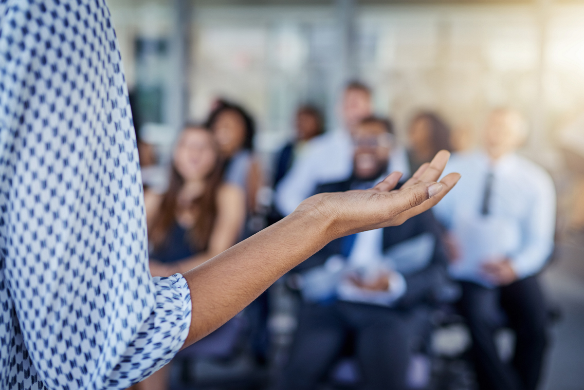 Close up image of a person's arm extended, as if expressing themselves while speaking. They stand in front of rows of people who are seated.