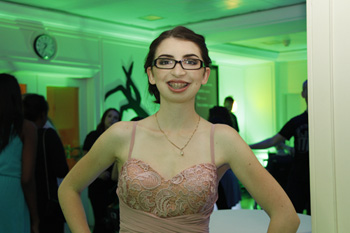A teen girl dressed in formal wear smiling. Behind her there are colourful lights and silhouettes of people dancing