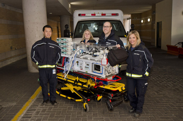 Four people in matching coats and pants stand by an incubator with an ambulance behind them.