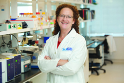 Woman stands in a lab wearing a lab coat.