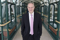 Man stands in SickKids atrium wearing a suit and pink tie with hands in pockets.