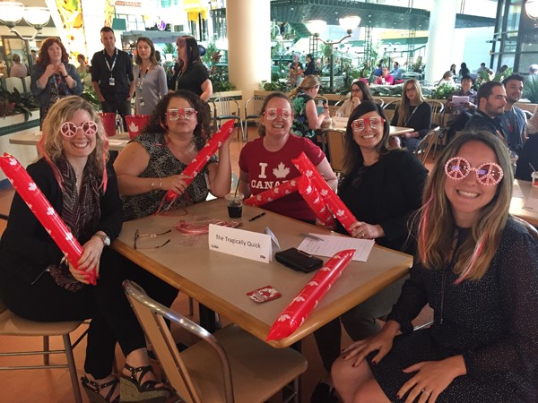 Five women seated around a table each wearing glittery dress up glasses.