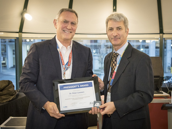 Two men stand together wearing suit jackets. One holds a plaque.