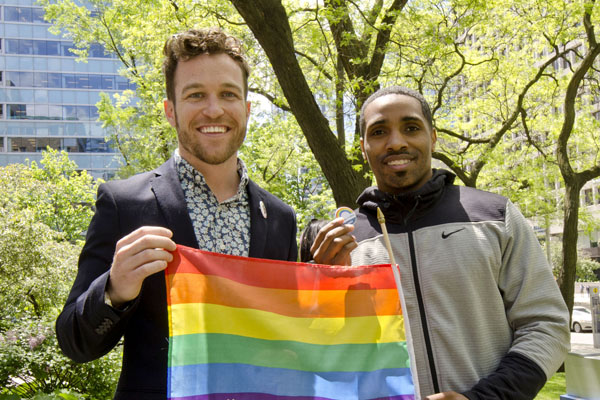 Two men stand side by side, one holds a rainbow flag.