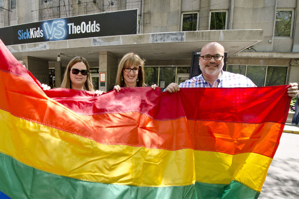 Three people stand together behind a very large rainbow flag.