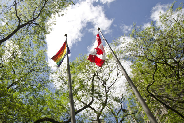 Rainbow pride flag and Canadian flag each flying from a flag pole. Photo taken from below.