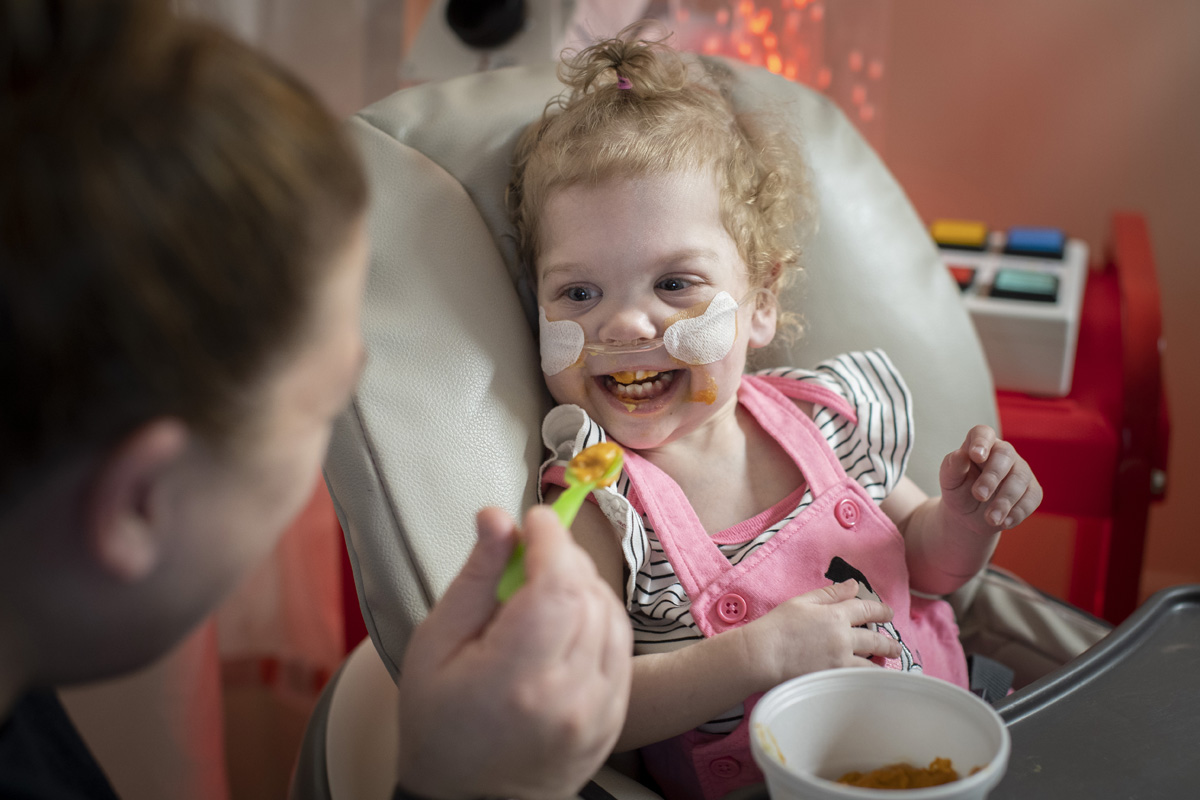 Toddler sitting in a high chair smiling at the adult who is feeding them with a spoon.