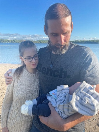 Man has arm around teen and holds a baby. The teen and man both look at the baby.