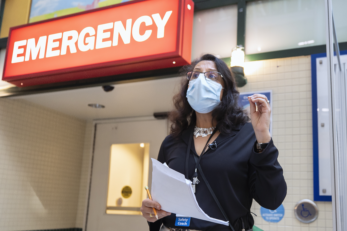 Person stands in front of Emergency department sign, wearing a mask and holding papers.