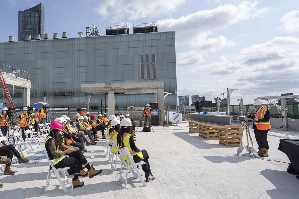 A man in construction gear stands at a podium speaking, while the audience, also wearing construction gear, sits in rows of chairs facing him