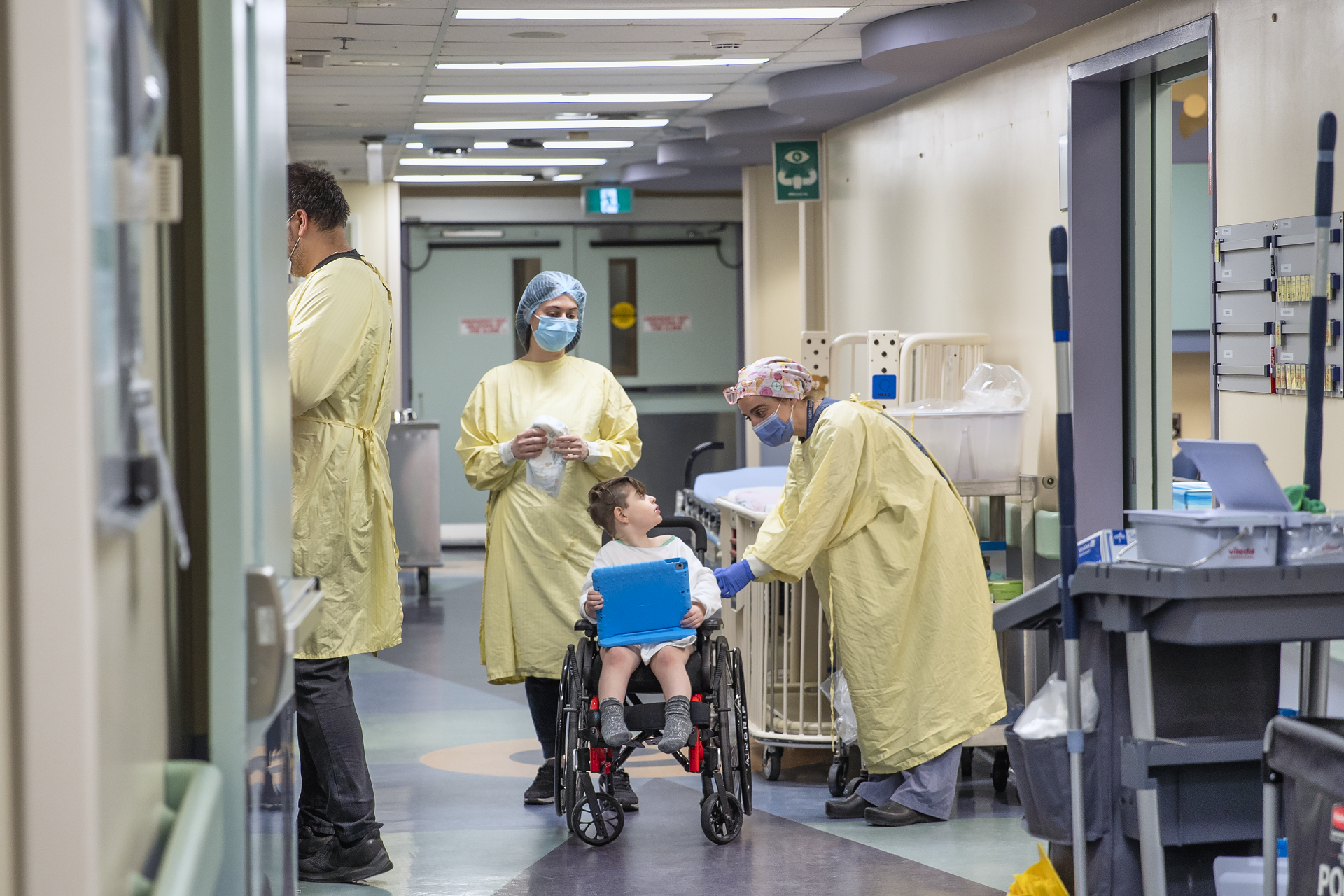 Child sits in a wheelchair holding a tablet. Three adults in medical gowns, caps and masks are present. One leans over and speaks to the child.
