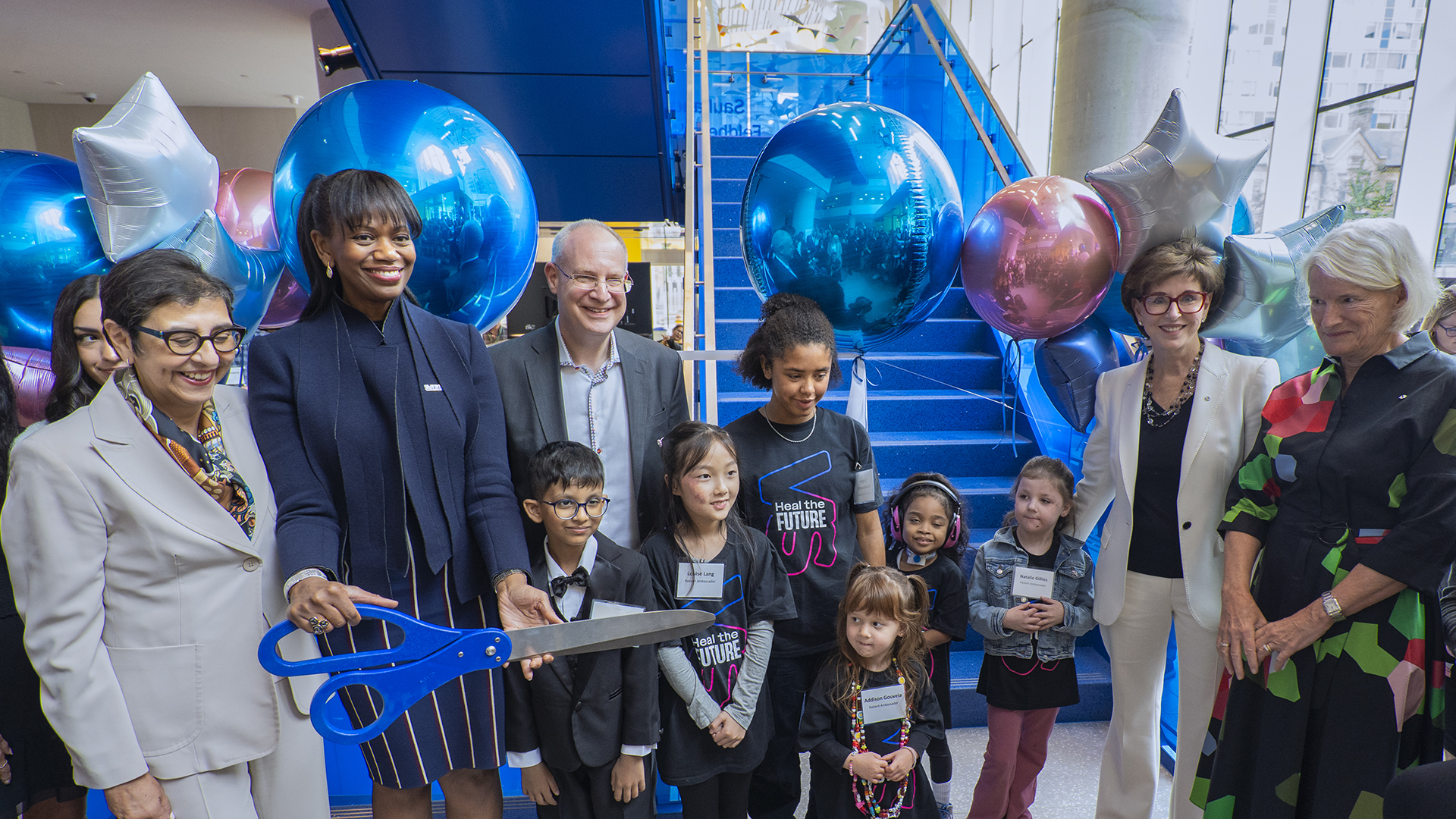 A group of children and adults, including one adult holding a large pair of scissors, gathered around a staircase that is roped off with a large ribbon and balloons