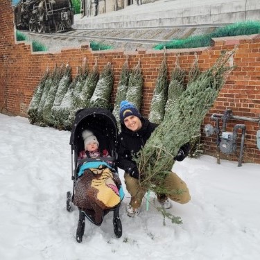 Brett holds a Christmas tree, kneeling beside Jeff, who is in his stroller.