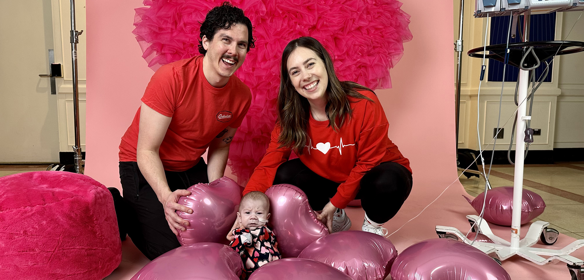 A man and a woman dressed in red T-shirts posing with their baby in front of a heart backdrop and pink heart balloons on the floor.